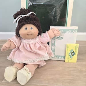 May include: A Cabbage Patch Kids doll with dark brown yarn hair tied with a white ribbon, wearing a light pink dress with white lace trim and cream-colored shoes. The doll is accompanied by its original box, a paper document, and a yellow card.