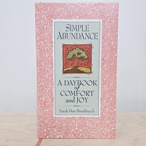 May include: A hardcover book titled "Simple Abundance: A Daybook of Comfort and Joy" by Sarah Ban Breathnach. The book has a pink floral patterned cover with a rectangular frame. The book's cover features a tree illustration.