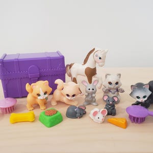 Puede incluir: Un cofre de juguetes morado con una variedad de animales de juguete, incluyendo un caballo, gatos y ratones. También hay herramientas de aseo para juguetes, como cepillos y un peine.