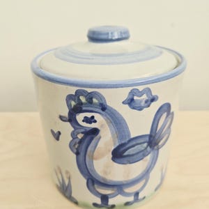 Puede incluir: Un tarro de cerámica con tapa, decorado con un diseño de gallo pintado a mano en azul sobre un fondo color crema. El tarro incluye detalles pintados en azul de un pequeño corazón, una forma de labios y hierba estilizada en la base, con borde azul.