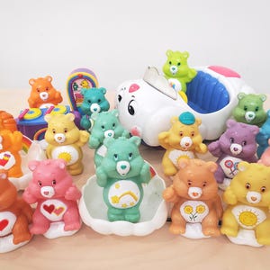 Könnte beinhalten: Eine Sammlung farbenfroher Care Bear-Figuren, darunter orange, gelbe, rosa, grüne und blaue Bären. Einige Bären befinden sich in weißen, muschelförmigen Behältern. Ein weißes Spielzeugauto mit blauen Akzenten ist ebenfalls vorhanden.