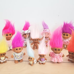 Puede incluir: Una colección de muñecos Troll vintage con pelo puntiagudo y colorido. Los muñecos tienen varios atuendos y accesorios, incluyendo vestidos, baberos y sombreros. Los colores del pelo incluyen rosa, naranja, amarillo, morado y blanco.
