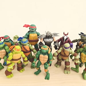 Può includere: Una collezione di action figure delle Tartarughe Ninja che include varie versioni delle quattro tartarughe con le loro caratteristiche maschere colorate, insieme a un personaggio di Shredder in armatura argentata e uno di Splinter in tunica viola.