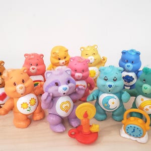 May include: A collection of colorful Care Bear figurines, each with unique colors and belly badges. The bears are in various poses, with a few accessories like a candle holder and a clock. The toys are made of plastic and are arranged on a light-colored surface.