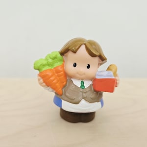 May include: A small plastic figurine of a person with brown hair, wearing a tan vest over a white shirt with a green tie, a white apron, and blue pants. The figure holds a bunch of orange carrots in one hand and a red basket in the other.
