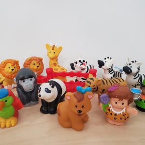 May include: A selection of colourful plastic toy animals and figures, including lions, a giraffe, zebras, a tiger, a panda, a bear, a parrot, a gorilla, and two human figures. The toys are arranged on a light brown surface.