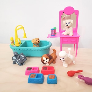 Puede incluir: Un juego de aseo para mascotas con una bañera turquesa con un grifo amarillo, un tocador rosa con espejo y varios perros y gatos de juguete. Incluye comederos, cepillos y comida para mascotas.