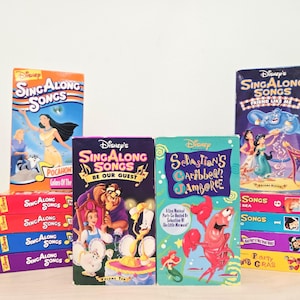 May include: A collection of Disney Sing Along Songs VHS tapes featuring various animated characters including Pocahontas, Belle, the Beast, Sebastian, and Aladdin. The stack includes titles like Be Our Guest and Sebastian's Caribbean Jamboree.