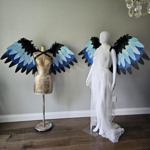 May include: Two sets of angel wings with a black and blue gradient design. The wings are displayed on a gold dress form and a white dress form. The wings have a feathered appearance and are attached to the dress forms.