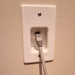 May include: A white plastic wall plate with a cutout for an ethernet cable. A gray ethernet cable is plugged into the wall plate.