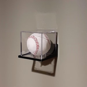 May include: A baseball in a clear acrylic display case, mounted on a black shelf. The baseball is white with red stitching. The case is fixed to a wall, creating a shadow.