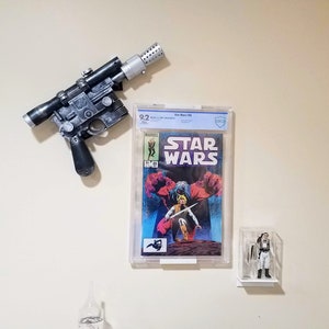 Puede incluir: Un cómic de Star Wars en un protector de plástico transparente, un bláster negro y plateado, y una figura de acción de Star Wars en una caja de plástico transparente en una pared blanca.