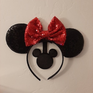 Puede incluir: Diadema de orejas de ratón negras con un lazo rojo de lentejuelas. La diadema tiene una silueta de Mickey Mouse de fieltro negro.