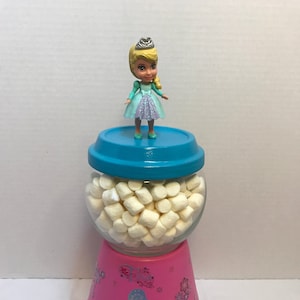 May include: A pink ceramic gumball dispenser with a clear glass jar filled with white marshmallows. A small plastic doll wearing a blue dress and a crown is on top of the dispenser lid.
