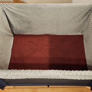May include: A rectangular pet bed with a dark blue frame. The interior features a burgundy plush base and is lined with a light gray, textured fabric. The bed is designed for comfort and support.