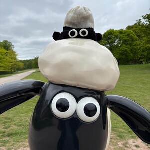 Könnte beinhalten: Eine schwarz-weiße Schafstatue mit einer kleinen Strickmütze. Das Schaf hat große, weiße Augen und eine glänzende Oberfläche. Die Mütze ist grau und weiß mit einem schwarzen Rand und zwei weißen Augendetails.