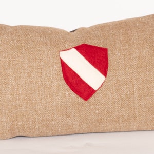 Puede incluir: Cojín rectangular beige con un escudo rojo y blanco. El cojín tiene una tela texturizada y un reverso oscuro. El escudo es de color rojo brillante con una franja blanca en el centro.
