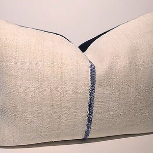 Puede incluir: Una almohada de lino blanco con una franja azul en el centro. La almohada es rectangular y tiene una superficie ligeramente texturizada.