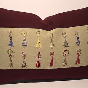 Puede incluir: Cojín decorativo rectangular con borde y reverso burdeos. El panel central es de color beige claro con un patrón repetido de figuras estilizadas que llevan cestas. Las figuras son de varios colores, incluyendo azul, amarillo y rojo.