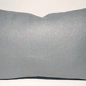 Puede incluir: Una almohada de tiro gris claro con un brillo sutil. La almohada está hecha de una tela suave y texturizada.