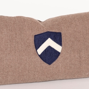 Puede incluir: Una almohada de tiro marrón con un diseño de escudo bordado en azul marino y blanco.