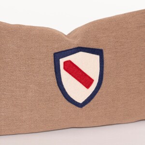 Puede incluir: Una almohada rectangular marrón con un diseño de escudo bordado en blanco y rojo. El escudo tiene un contorno azul marino.