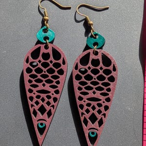 May include: A pair of dangle earrings with a teardrop shape, featuring a burgundy laser-cut design. The earrings have teal accents and gold-colored ear wires. A pink measuring tape is next to the earrings.