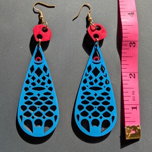 Può includere: Un paio di orecchini blu a goccia con un design geometrico ritagliato, top circolari rossi e ganci color oro. Gli orecchini sono esposti accanto a un metro a nastro rosa.