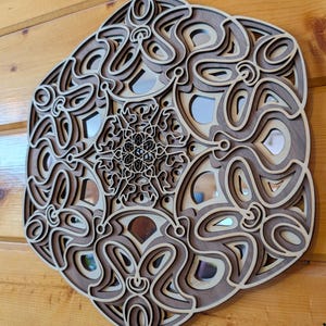 Può includere: Un'opera d'arte da parete in legno a strati con un design complesso e simmetrico. L'opera presenta un mix di tonalità marroni chiare e scure, con accenti a specchio. L'intricato motivo ricorda un mandala o un ornamento decorativo.