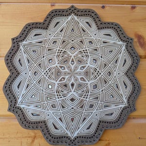 May include: A layered wooden mandala with a complex geometric design. The artwork features a central star-like pattern surrounded by concentric layers of geometric shapes in shades of brown and white. The outer edge has a dotted border.