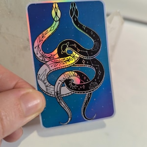 Adesivo místico celestial de duas cobras impresso em papel holográfico prateado, proporcionando um efeito arco-íris dependendo da luz.