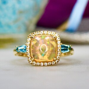 May include: A gold ring with a large, iridescent opal gemstone surrounded by a halo of small diamonds. Two smaller, blue gemstones are set on either side of the opal.