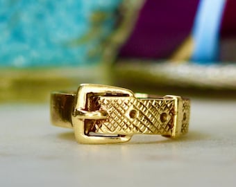 May include: A gold-colored ring in the shape of a belt buckle. The ring has a textured band and buckle, with a classic design. The ring is set against a blurred background.