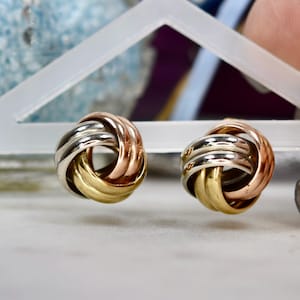 May include: A pair of tri-color earrings. Each earring features a knot design with interwoven bands of gold, silver, and rose gold. The earrings are displayed on a white surface, with a blurred background.