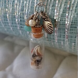 May include: A silver chain necklace with a small glass bottle charm. The bottle is filled with sand and seashells. The necklace also has a small silver seashell charm and a pearl charm.