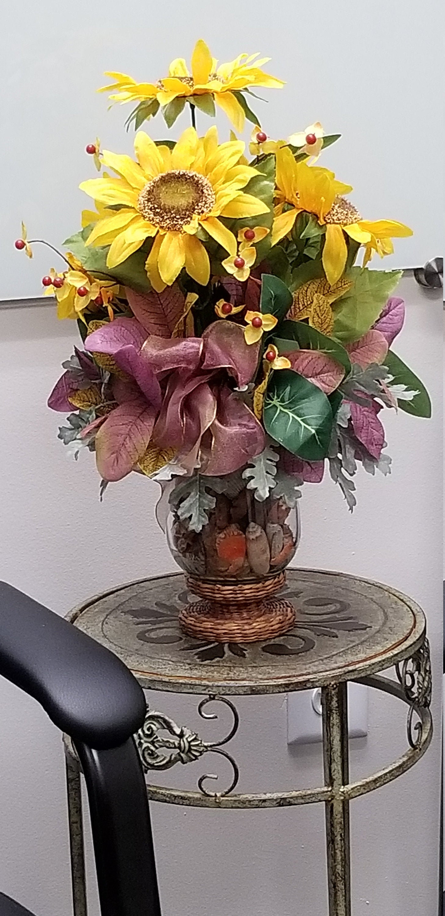 May include: A faux sunflower arrangement in a clear glass vase with a wicker base. The vase is filled with brown and orange decorative stones. The arrangement is on a round, metal table with a distressed finish.
