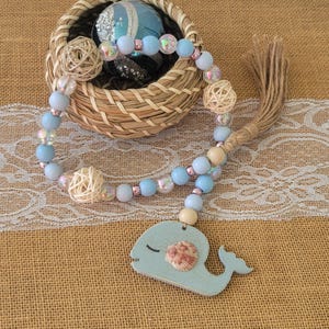 May include: A decorative garland featuring light blue and iridescent beads, wooden beads, and woven spheres. A light blue whale-shaped pendant with a seashell accent hangs from the garland. The garland is displayed with a woven basket and a jute tassel.