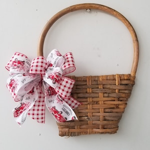 Pode incluir: Uma cesta de vime castanho com uma pega, decorada com um laço de xadrez vermelho e branco. O laço tem uma fita branca com um camião vermelho e o texto "Country Farmhouse" impresso nele.