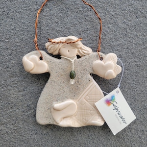Puede incluir: Un adorno de ángel de cerámica con un colgador de alambre. El ángel lleva un vestido blanco con un collar de cuentas verdes y tiene un halo. El adorno está hecho por Edgewater Designs.