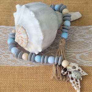 Pode incluir: Uma guirlanda decorativa de contas com contas de madeira cinza, azul claro e creme. A guirlanda é acentuada com uma borla de juta e pingentes de concha, perto de uma grande concha branca.