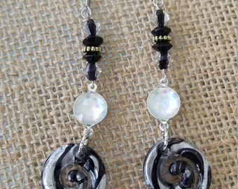May include: A pair of silver earrings with black and white swirl designs. The earrings feature a silver hook, a clear iridescent bead, and black beads with gold accents.