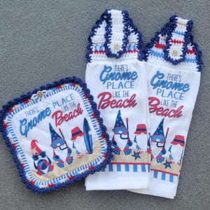 May include: A set of kitchen textiles with a beach theme. Includes two white hand towels and a square pot holder, all with a blue, red, and white gnome design and the text "There's Gnome Place Like the Beach."