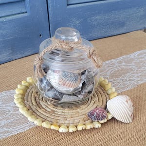 May include: A glass jar filled with seashells, secured with twine, rests on a woven mat adorned with small, pale-yellow shells. Two seashells are placed next to the jar. The background features blue shutters and a burlap surface.