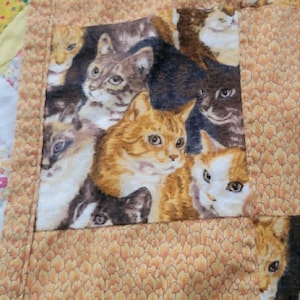 Op de afbeelding: Een patchworkquilt met een herhalend patroon van kattengezichten in verschillende kleuren, waaronder oranje, bruin en wit. De quilt heeft een getextureerde, bruinachtige rand.