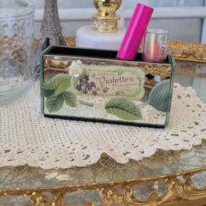 May include: A decorative black and mirrored vanity organiser with a floral and botanical design. The organiser holds a pink lipstick and a small glass container. The text "Violettes Francaises" is visible on the front.