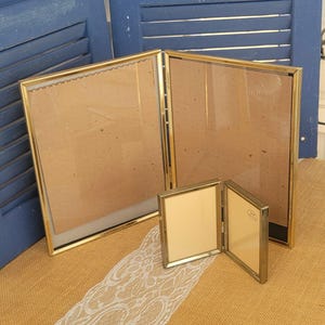 May include: A collection of gold-colored picture frames in various sizes. The frames are displayed on a tan surface with a blue shutter background. The frames have a classic, elegant design, suitable for displaying photographs or artwork.