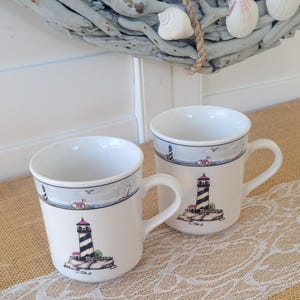 May include: Two white ceramic mugs with a lighthouse design. The mugs feature a black and white striped lighthouse with a red top, set against a coastal scene. The mugs have a handle and a blue border with a coastal design.