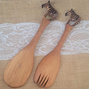 May include: A pair of wooden salad servers. One is a spoon, the other a fork. Each handle features a carved zebra head with black and brown stripes. The servers are light brown and rest on a burlap surface.