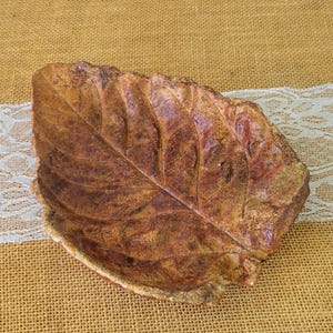 May include: A decorative leaf-shaped dish with a textured surface. The dish is primarily brown with reddish and gold accents, resembling a natural leaf. The dish is made of a sturdy material and is suitable for serving or display.