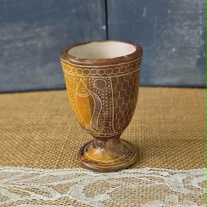 May include: A brown, hand-carved goblet with a fish design. The goblet has a wide rim, a detailed stem, and a base with circular patterns. The fish is etched in the center, with a honeycomb pattern on the sides. The interior is white.